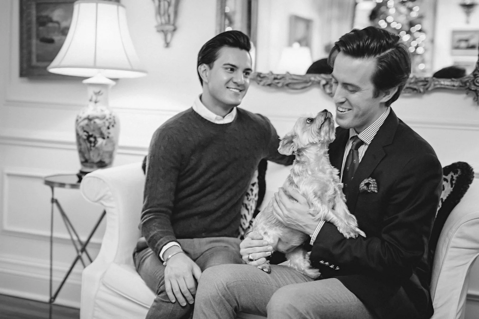 Two men smiling and looking at a small dog, which one of the men is holding, in an elegant living room with a lamp, a mirror, and holiday decorations.