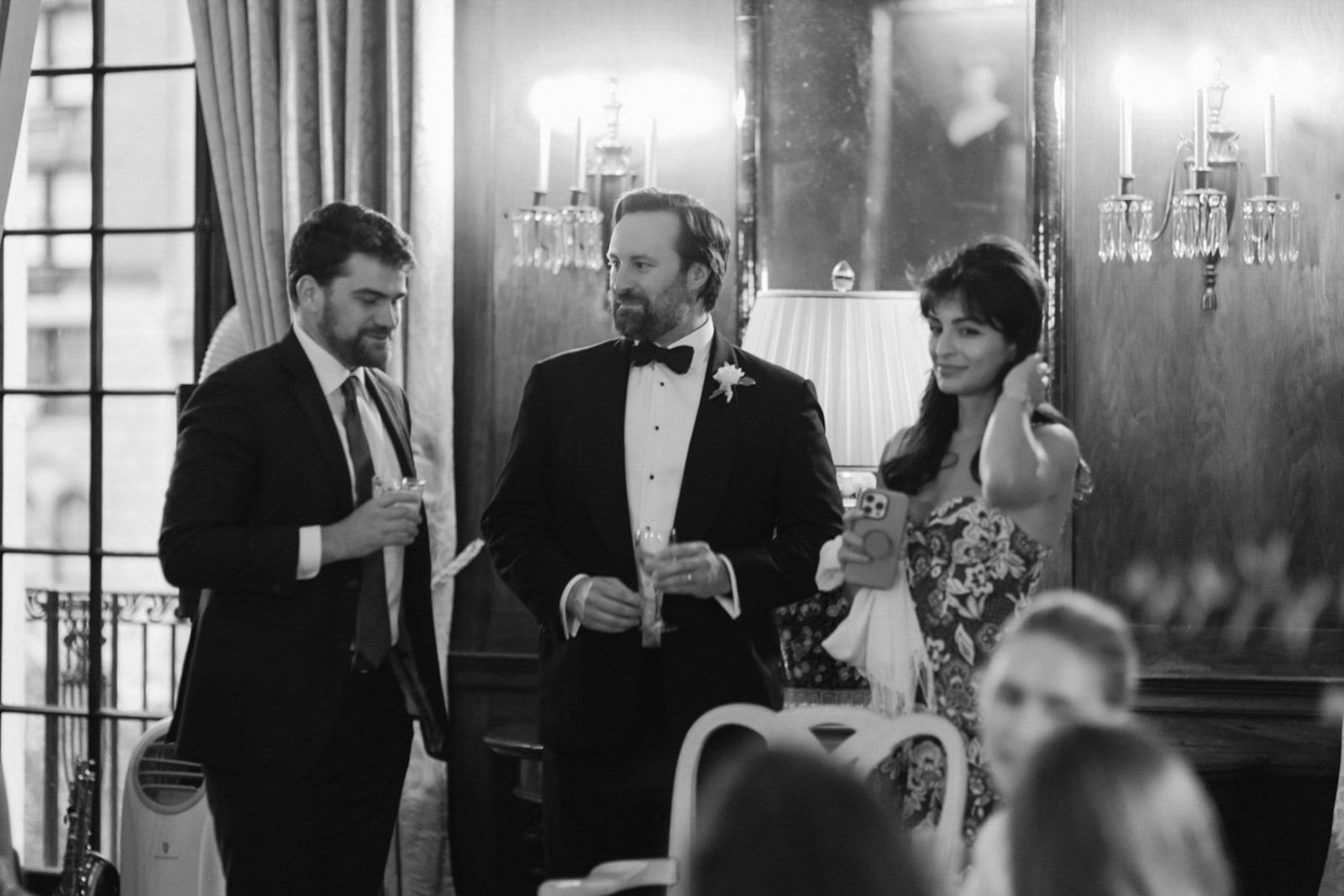 Black and white photo of three people at a formal event, two men in tuxedos and a woman in a dress, standing in a room with wooden wall panels, a large mirror, and candles.