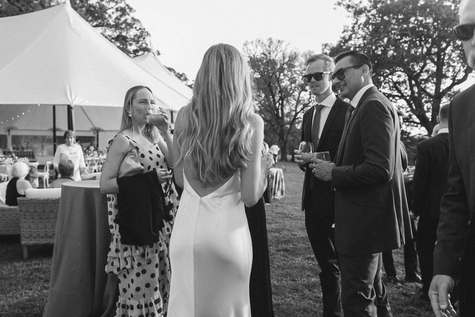 Black and white photo of a wedding reception gathering outdoors with several guests in formal attire, including a woman in a white gown talking to three men in suits, and a woman in a polka dot dress drinking from a glass.