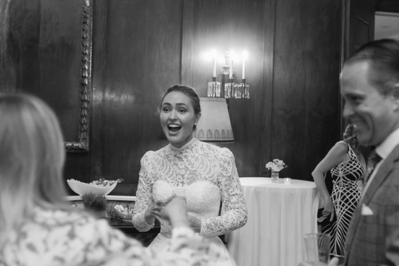 A black and white photo of a woman in a lace wedding dress smiling and talking at a formal event, with other people around her in an elegant setting with dark wood paneling and a lamp.