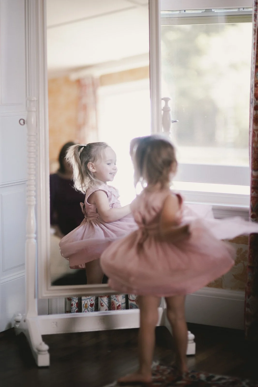 A young girl in a pink tutu is looking at her reflection in a full-length mirror, smiling as she admires herself. Sunlight streams through the window behind her, creating a soft, warm glow.