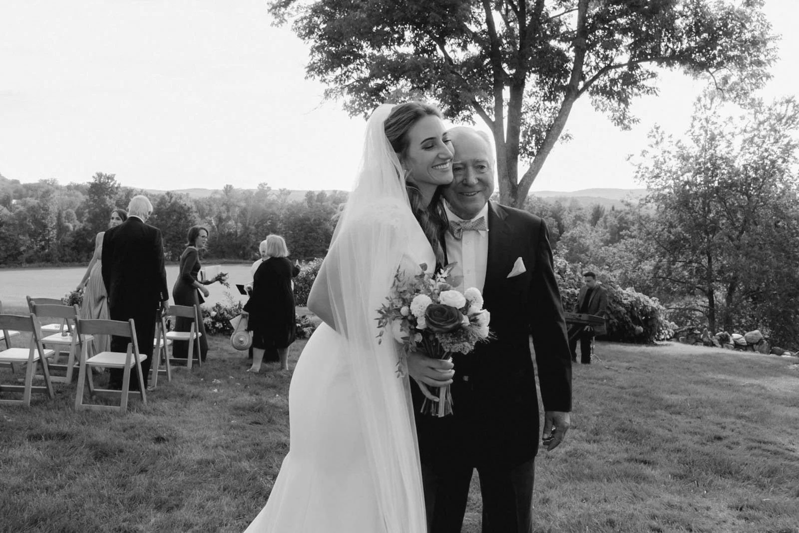A bride in a white wedding gown and veil, holding a bouquet of flowers, stands next to an older man in a tuxedo outdoors during a wedding ceremony, with trees and guests in the background.