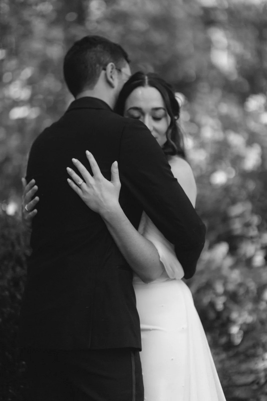 A black and white photo of a couple embracing outdoors, with the woman wearing a white dress and the man in a dark suit, their faces close and eyes closed.
