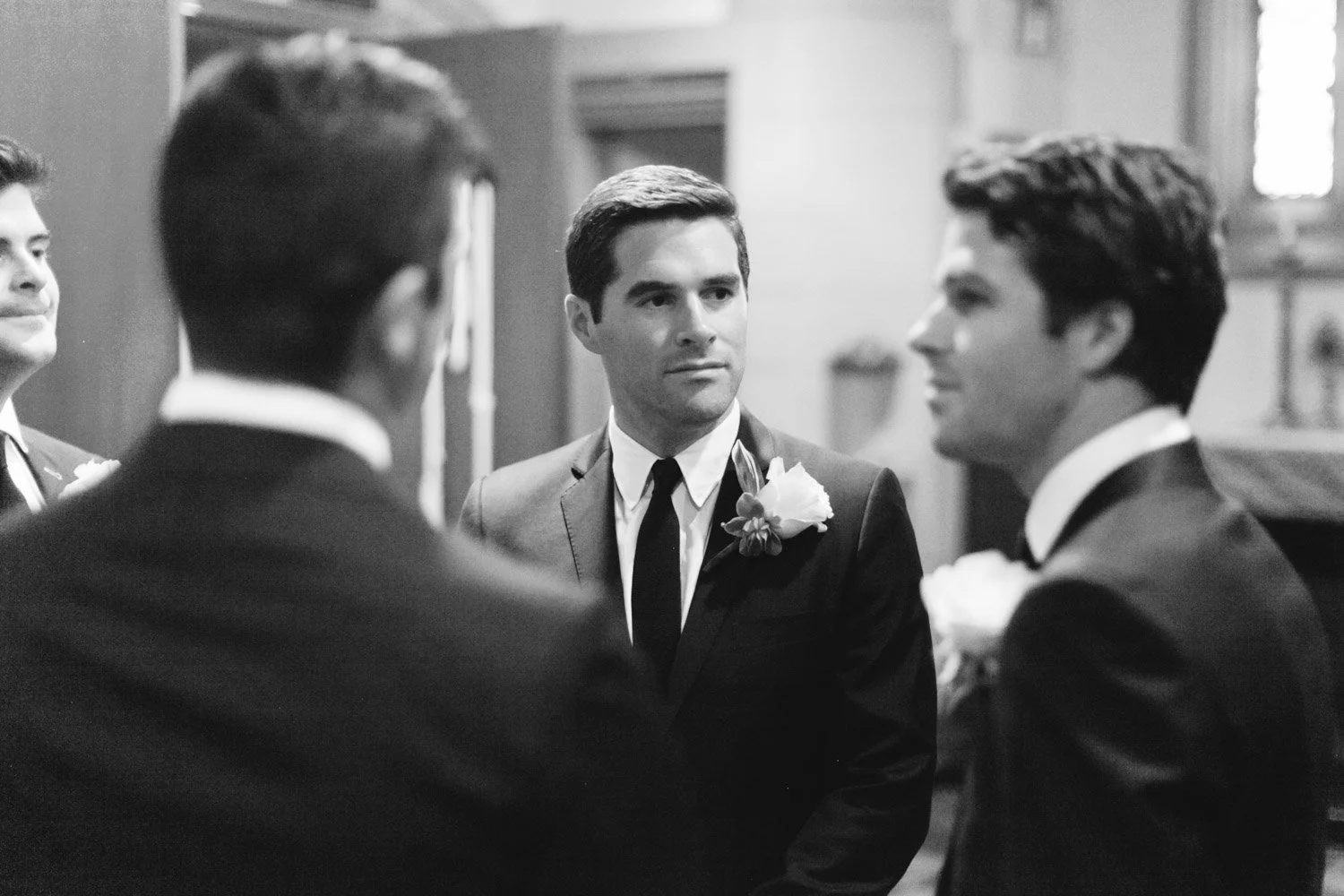 Black and white photo of four men in tuxedos at a wedding, with one man in focus looking at two men facing away from the camera.