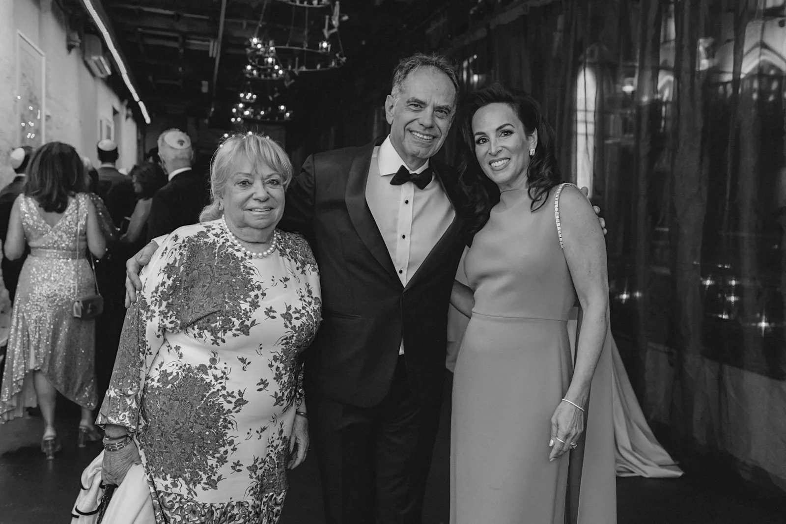 Three elegantly dressed people smiling at a formal event, with a woman in a floral dress, a man in a tuxedo, and another woman in a gown, in a decorated indoor venue.