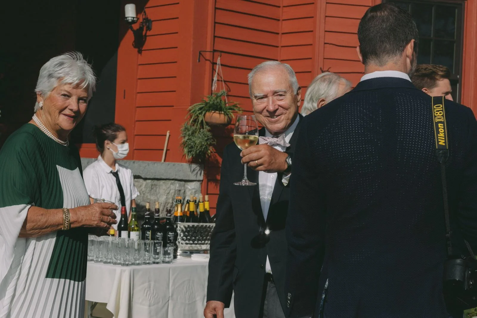 People at a social gathering, including a woman in a green dress, a man holding a wine glass, and others in the background, with bottles of wine and glasses on a table.