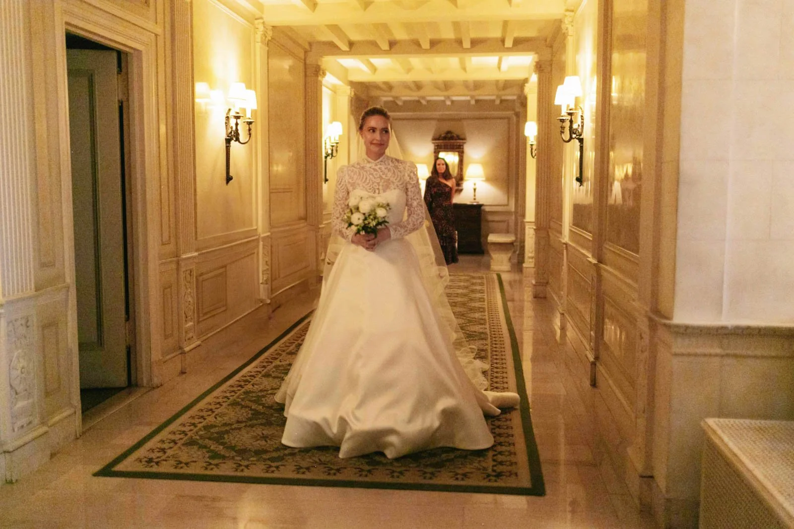 A bride in a white gown with lace details holding a bouquet of white roses, standing in a warmly lit, ornate hallway with wood-paneled walls and wall sconces, and a guest standing behind her.