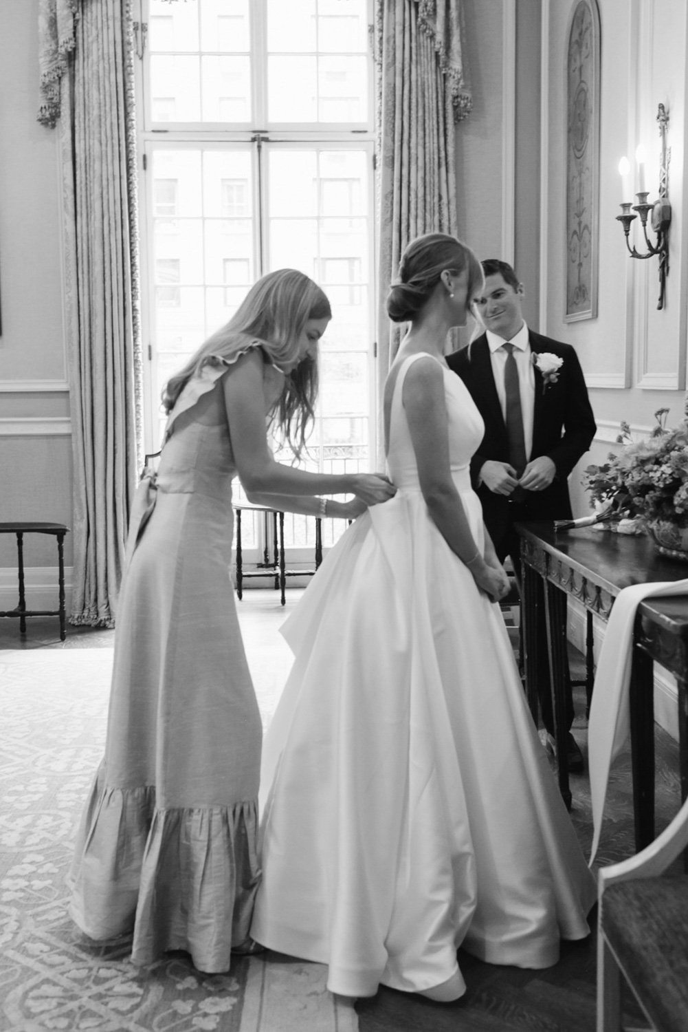 A black and white photo of a wedding ceremony with a bride and groom standing in a decorated room, a woman adjusting the bride's dress.