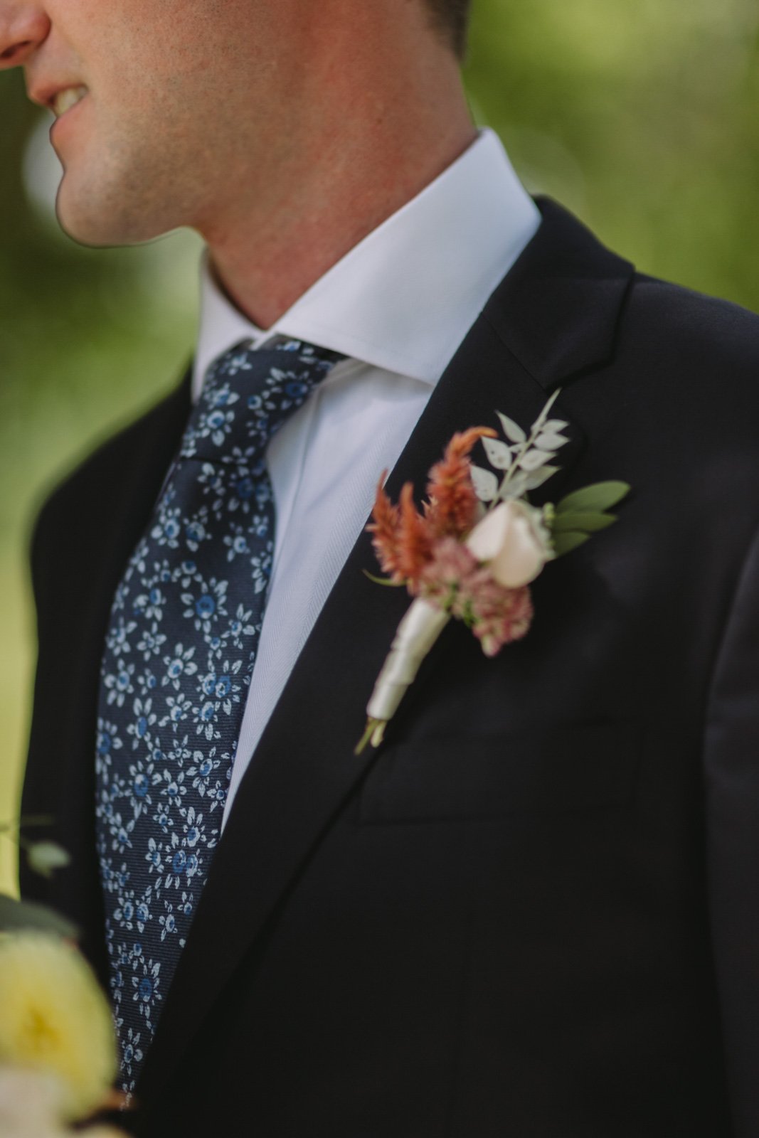 Close-up of a man in a black suit with a white shirt and a dark floral tie, wearing a boutonnière with pink and white flowers and greenery on his lapel, smiling outdoors.