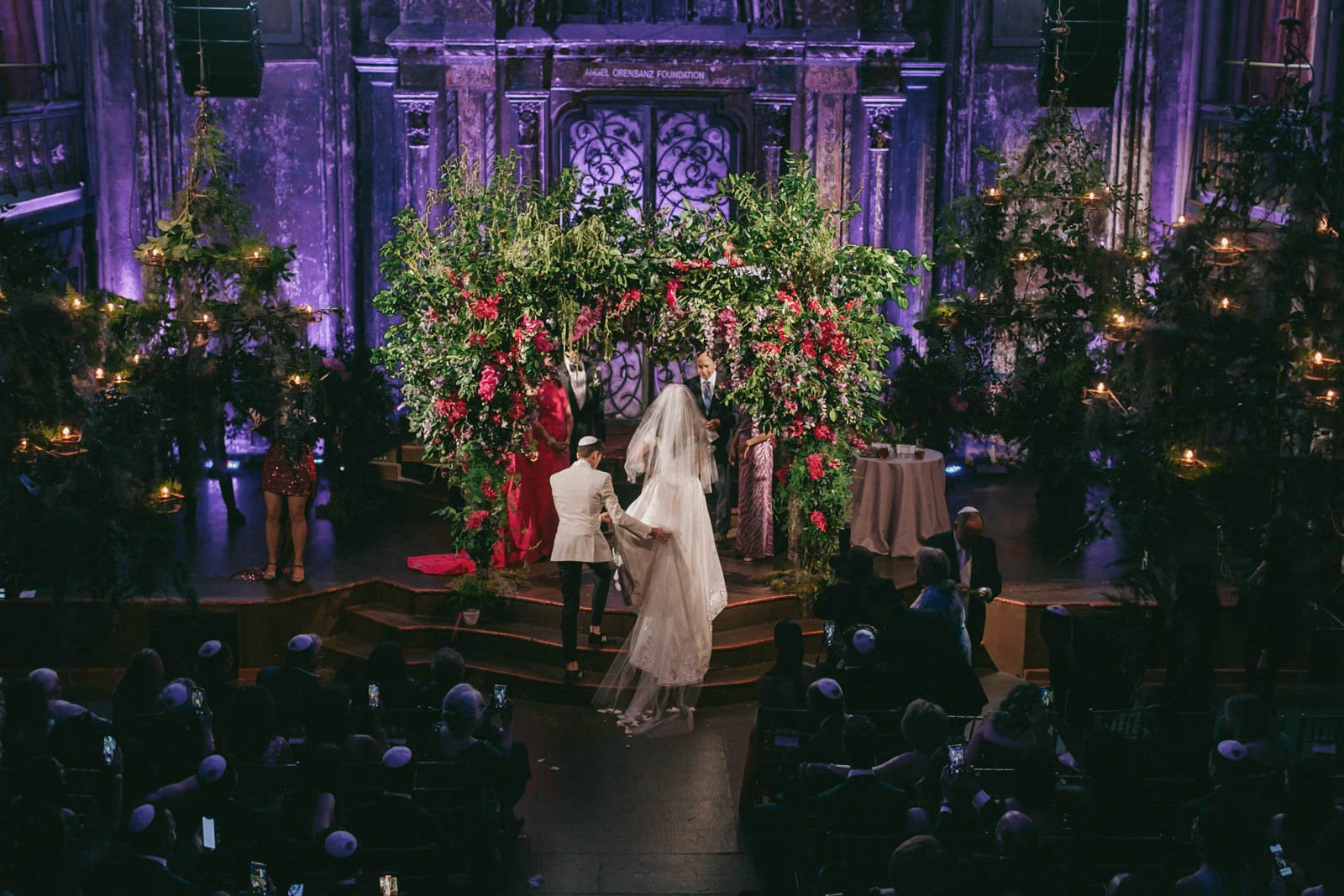 A wedding ceremony with a bride, groom, and officiant on stage, decorated with a colorful floral arch, in a grand hall with purple lighting and seated guests wearing kippahs.