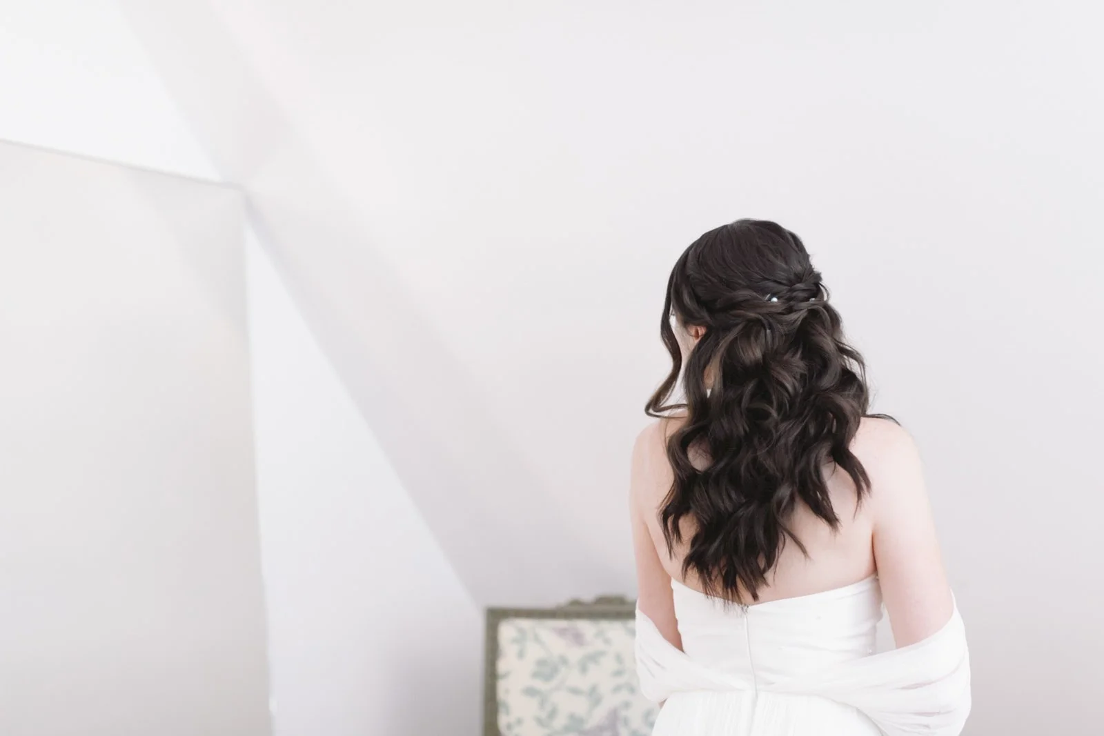 A woman with long dark wavy hair, wearing a strapless white dress, standing with her back to the camera in a bright, white room.