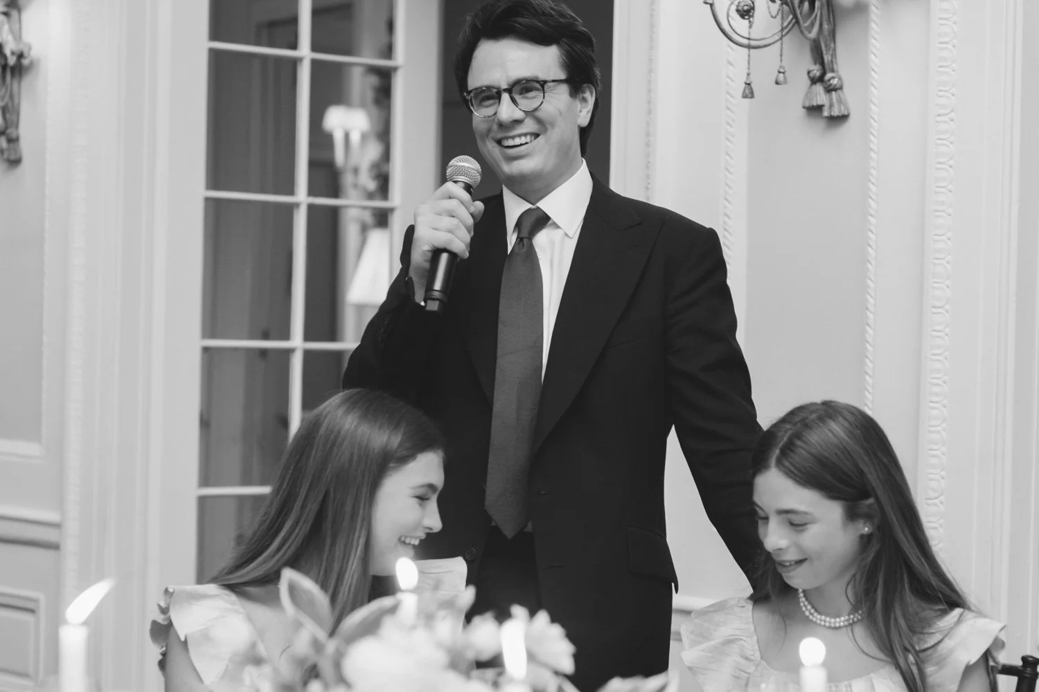 A man in a suit with glasses smiling and holding a microphone at a gathering, with two young girls sitting at a table, one smiling with closed eyes, and the other looking down, with candles and flowers on the table.