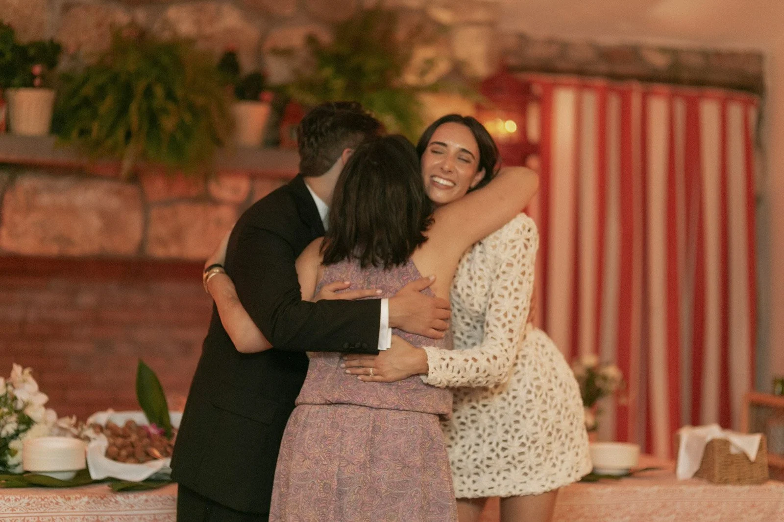 Three people hugging at a celebration or gathering, with warm lighting and decorated background.
