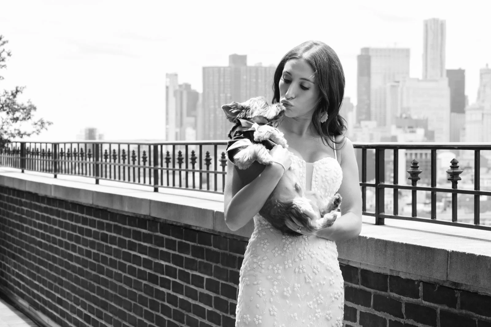 A woman in a dress holding and kissing a small dog on a rooftop with a city skyline in the background.