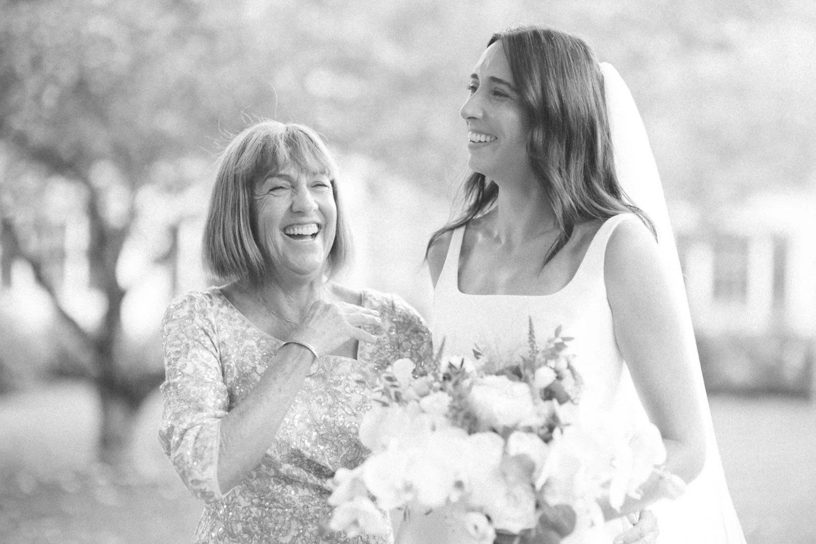 Two women, an older woman and a younger woman in a wedding dress, smiling and sharing a joyful moment outdoors, with the younger woman holding a bouquet of flowers.