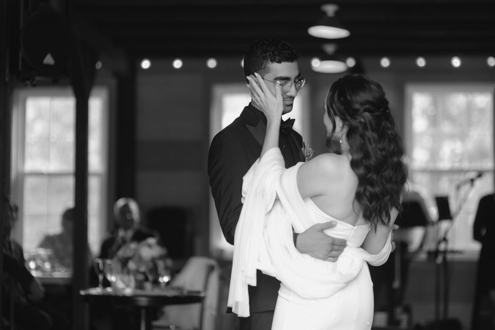 A couple dancing at their wedding reception, with the bride wearing a white dress and the groom in a tuxedo, in a dimly lit room with large windows.