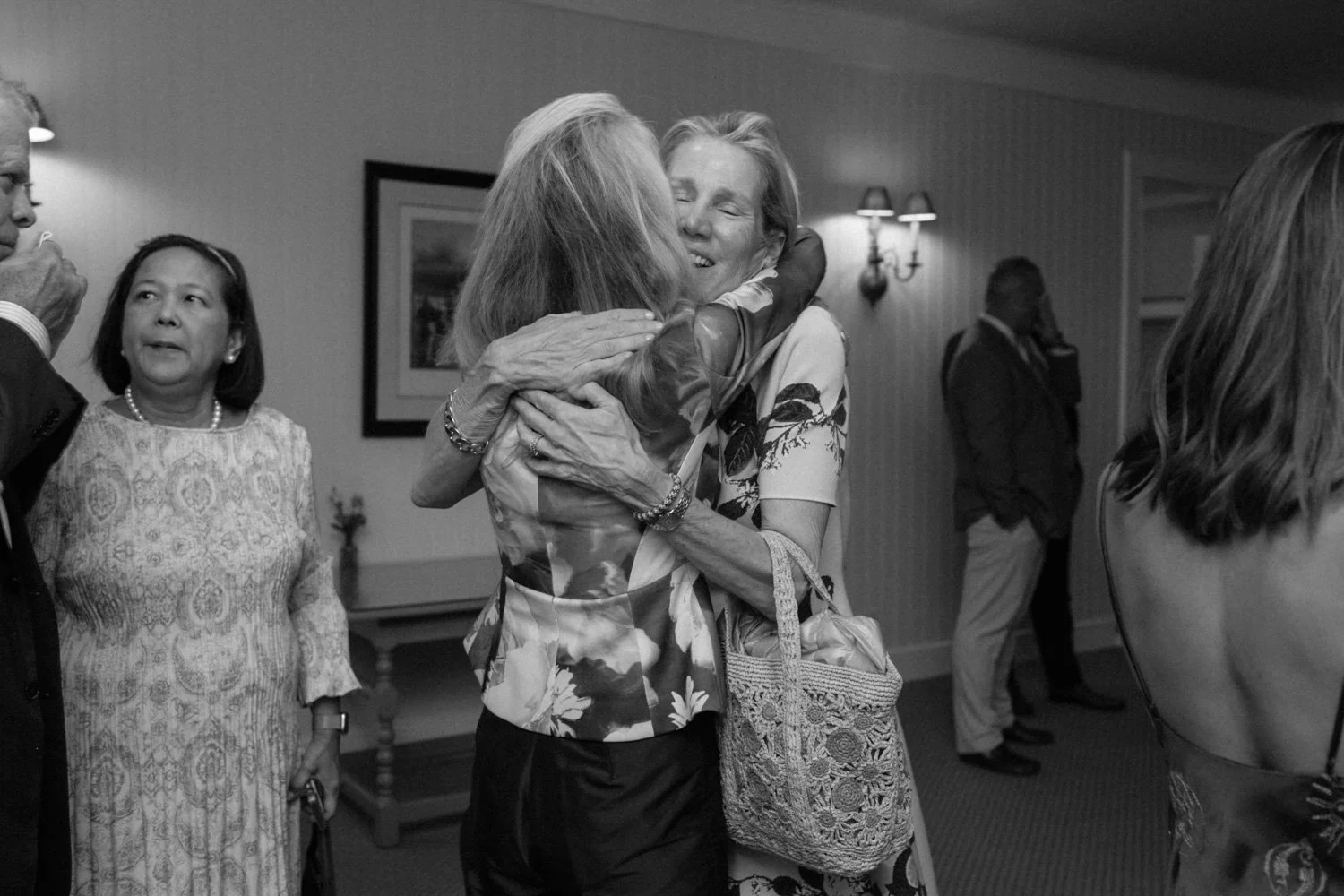 Two women embrace warmly at a social gathering, with other individuals in the background.