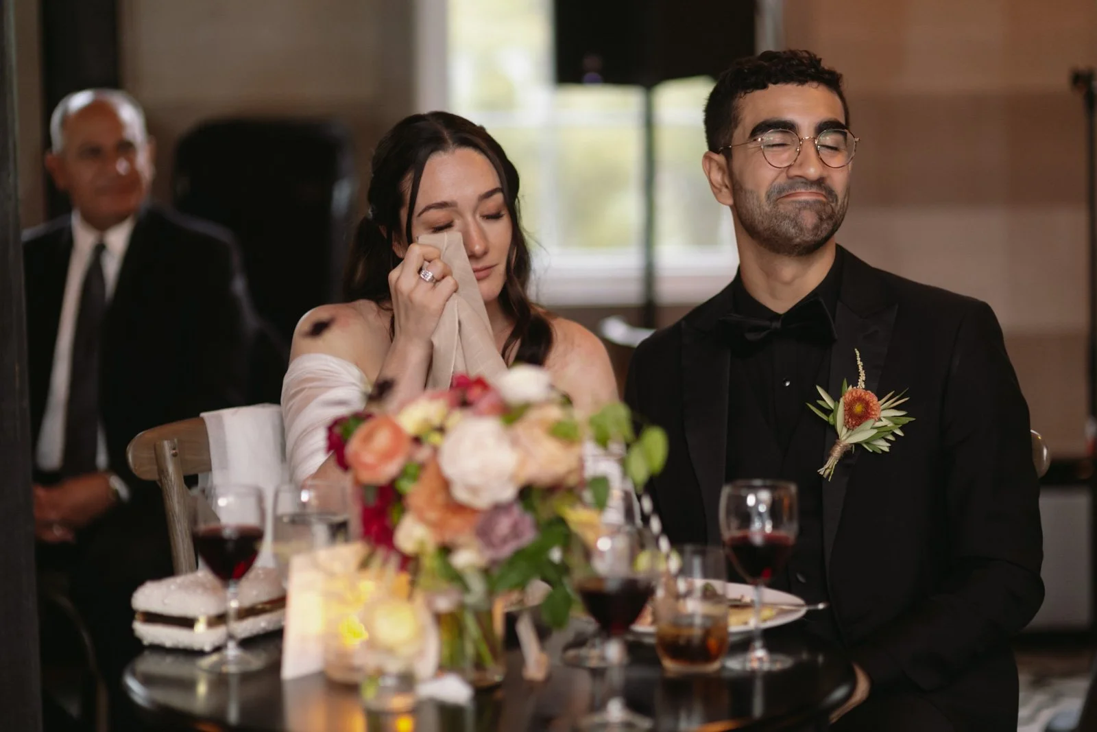 A woman at a wedding reception is crying and wiping her tears with a napkin, while a man next to her, dressed in a tuxedo, is smiling with eyes closed. There are glasses of wine, a floral centerpiece, and other guests in the background.