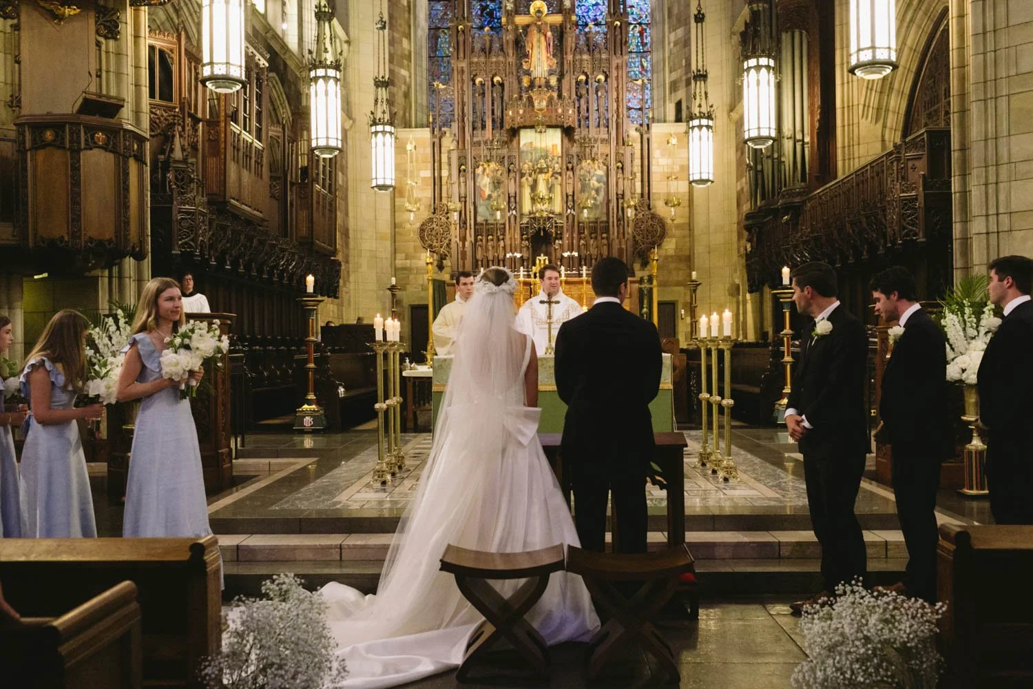 A wedding ceremony inside a church with a bride and groom standing at the altar, surrounded by bridesmaids and groomsmen. The church has tall stained glass windows and ornate wooden details.