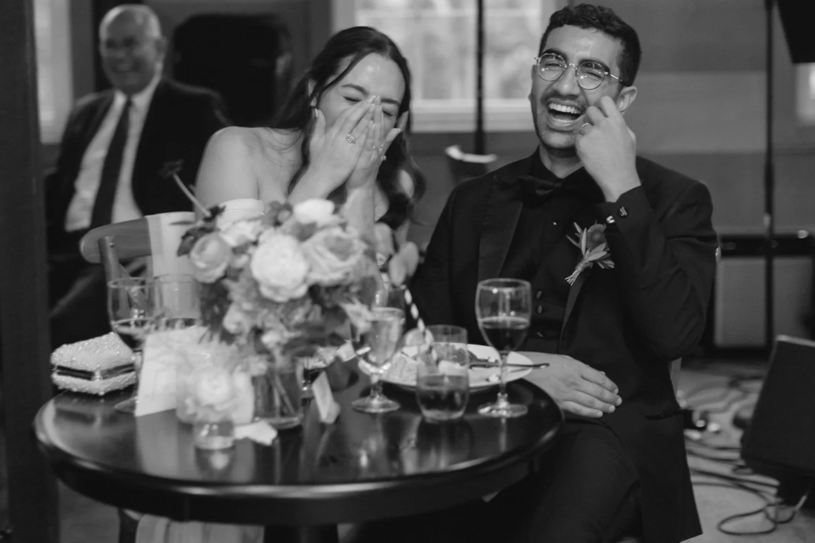 A wedding reception with a bride and groom laughing at a table with flowers and drinks, while a man in a suit smiles in the background.