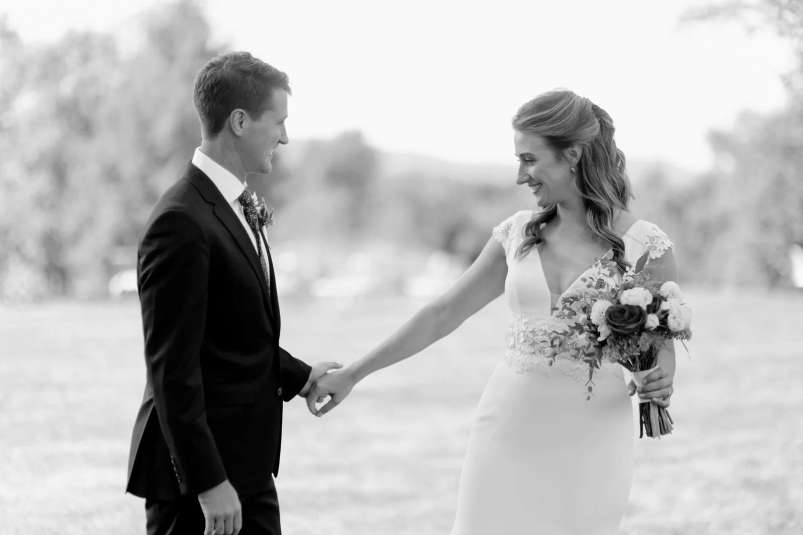 Black and white photograph of a bride and groom outdoors, holding hands and smiling at each other, the bride holding a bouquet of flowers.
