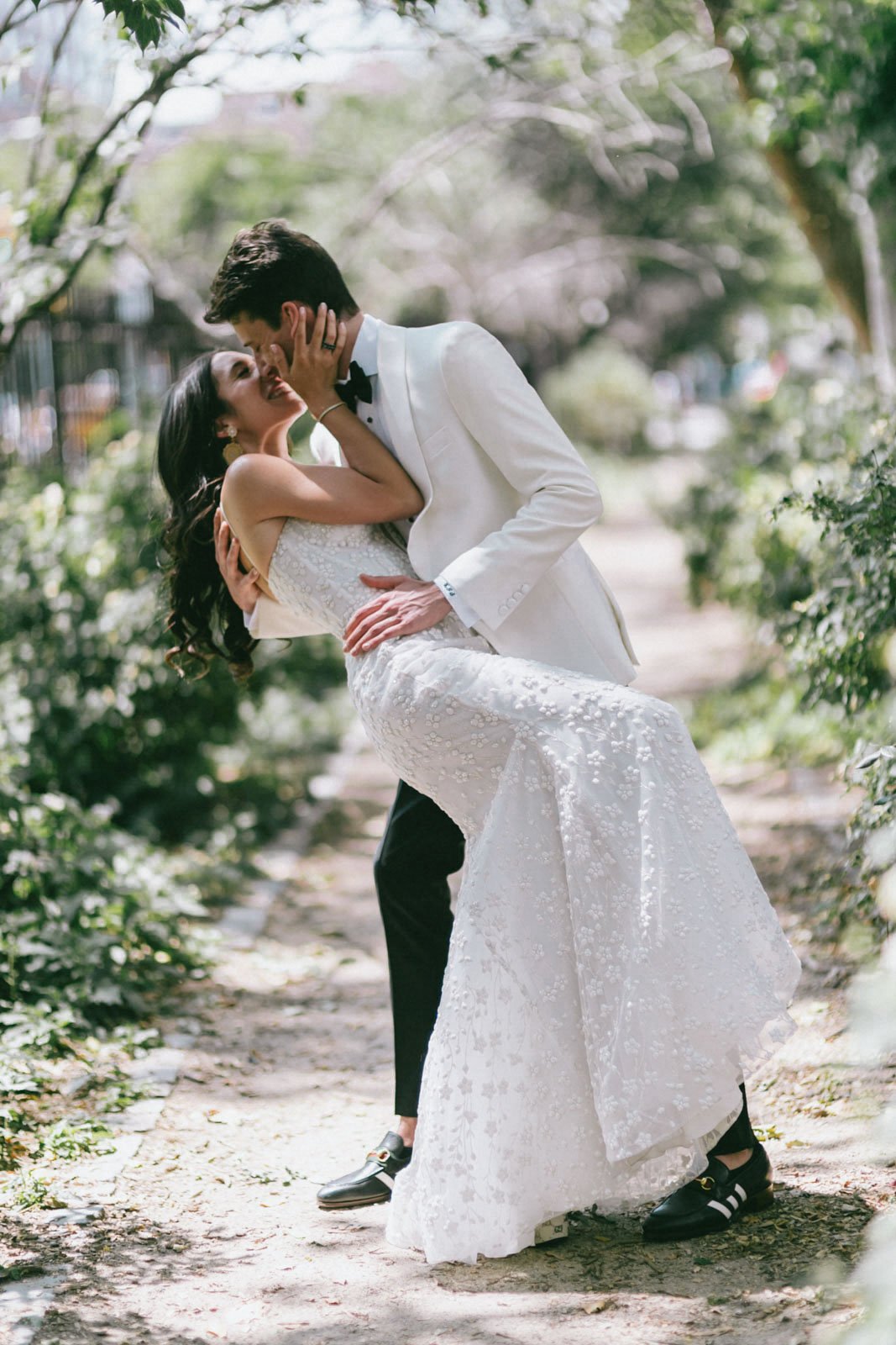 A bride and groom sharing an intimate moment outdoors, with the groom holding the bride as they lean in close to kiss, surrounded by greenery.