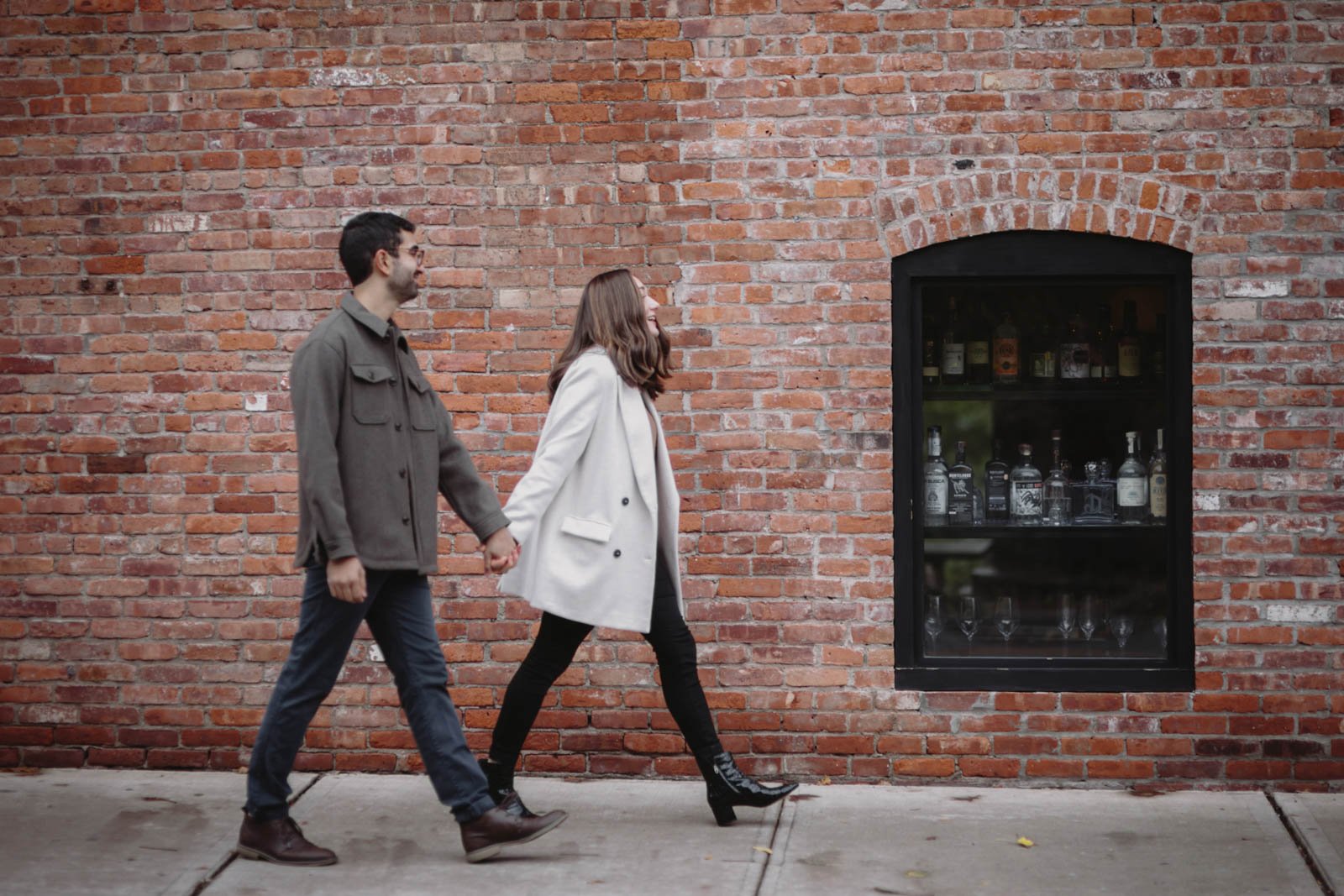 A man and woman holding hands and walking past a brick wall with a window display of liquor bottles.