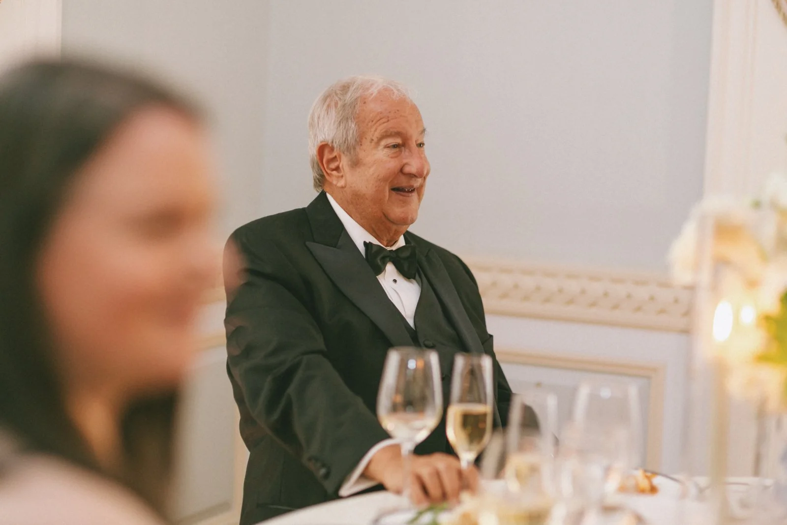 An elderly man in a tuxedo with a bow tie, smiling during a formal event, likely a wedding reception or banquet, with a blurred woman in the foreground and a decorated table with glasses in the background.