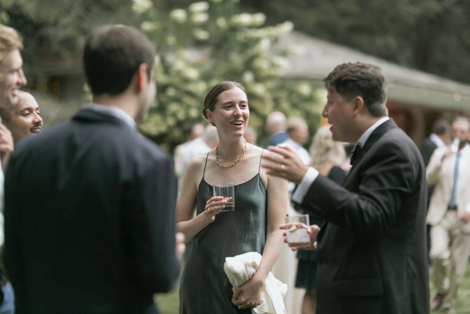 People at an outdoor gathering, including a woman in a black dress and gold jewelry, holding a glass, and a man in a black tuxedo talking to her, with other guests socializing in the background.