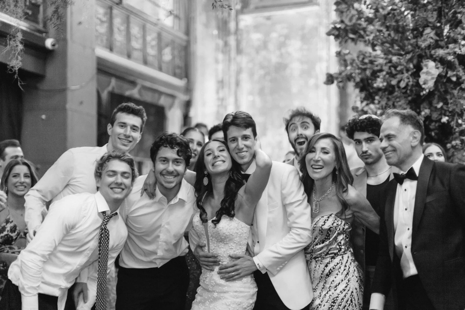 A group of people dressed in formal attire, celebrating at a wedding reception, with smiling faces and some taking selfies.
