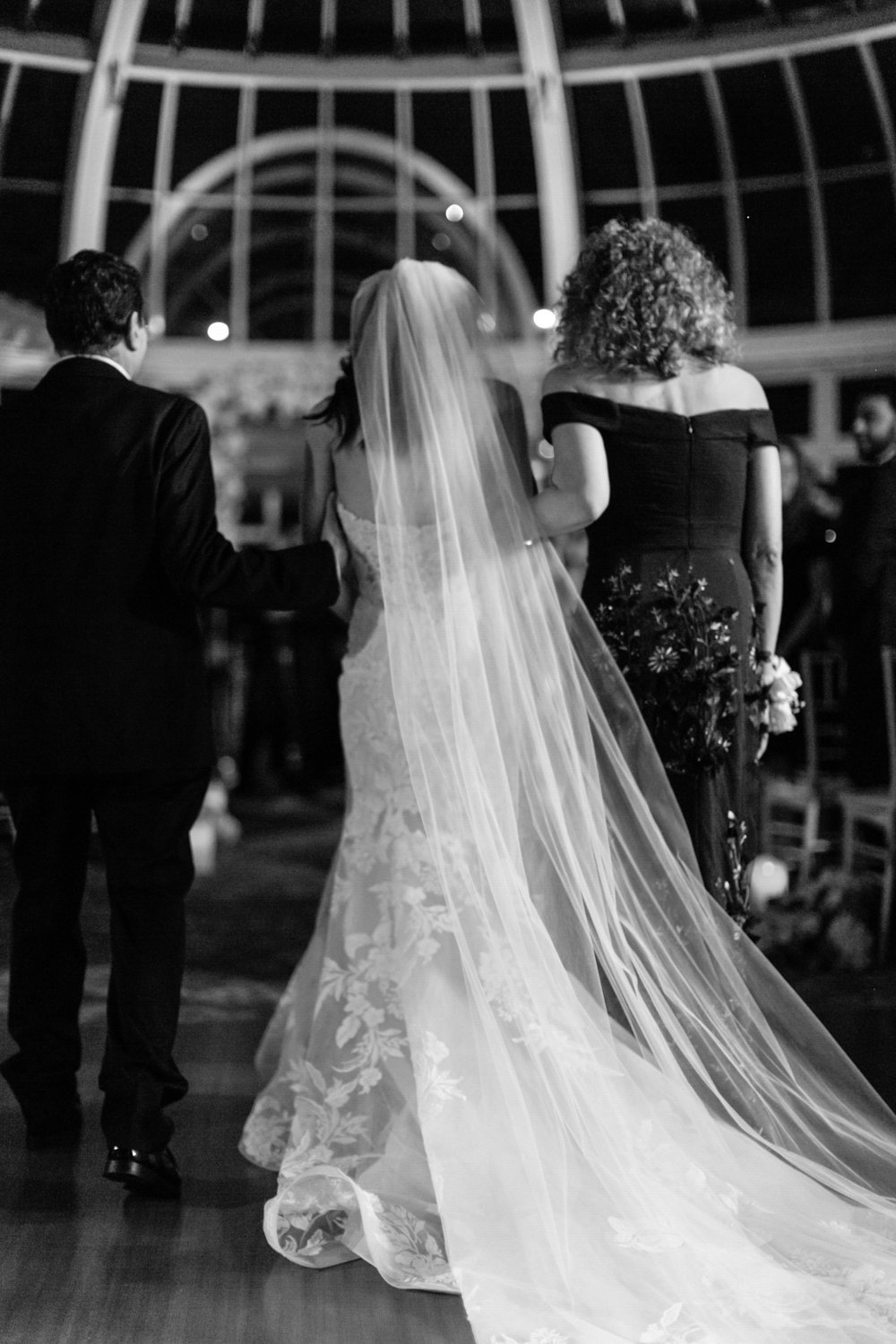Black and white photo of a bride in a wedding dress with a long veil, walking with two women, at a wedding reception or ceremony inside a venue with large windows and an arched ceiling.
