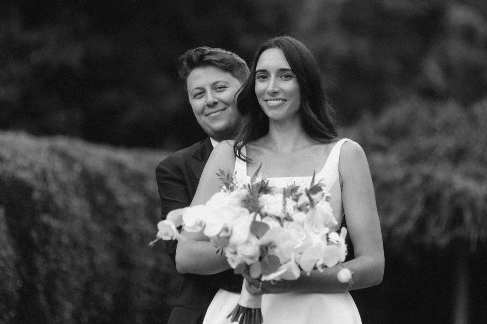 A black and white photo of a smiling couple, with the woman holding a bouquet of flowers, outdoors with trees in the background.