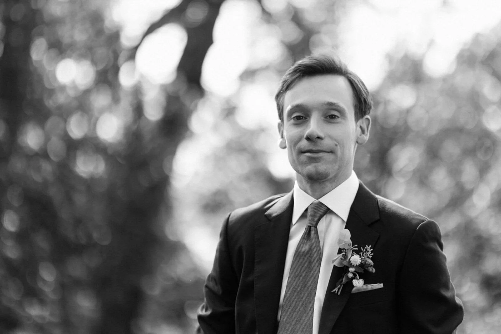 A black and white photo of a young man in a suit with a boutonniere, outdoors with blurred trees in the background.