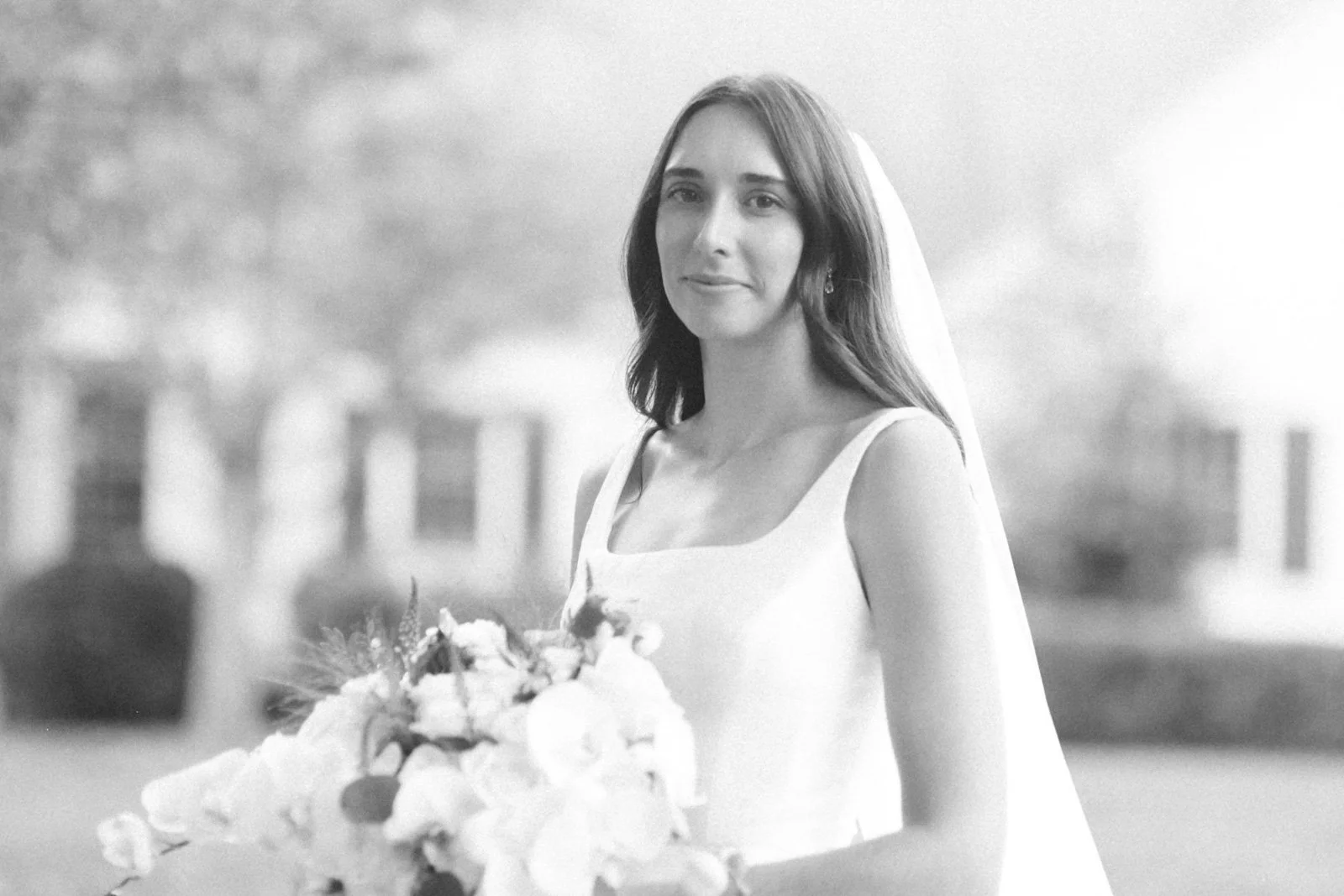 A woman in a white dress holding a bouquet, smiling softly, photographed outdoors in black and white.