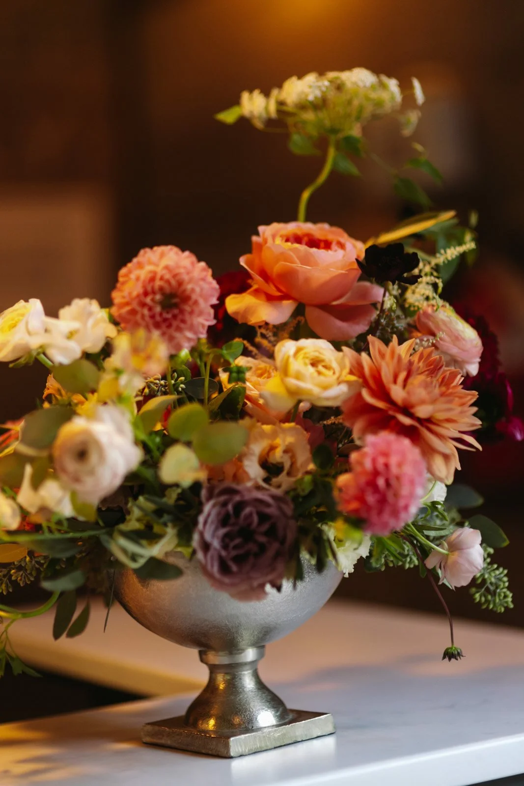 A silver pedestal bowl filled with pink, peach, and cream-colored flowers, including roses and dahlias, arranged on a white surface.