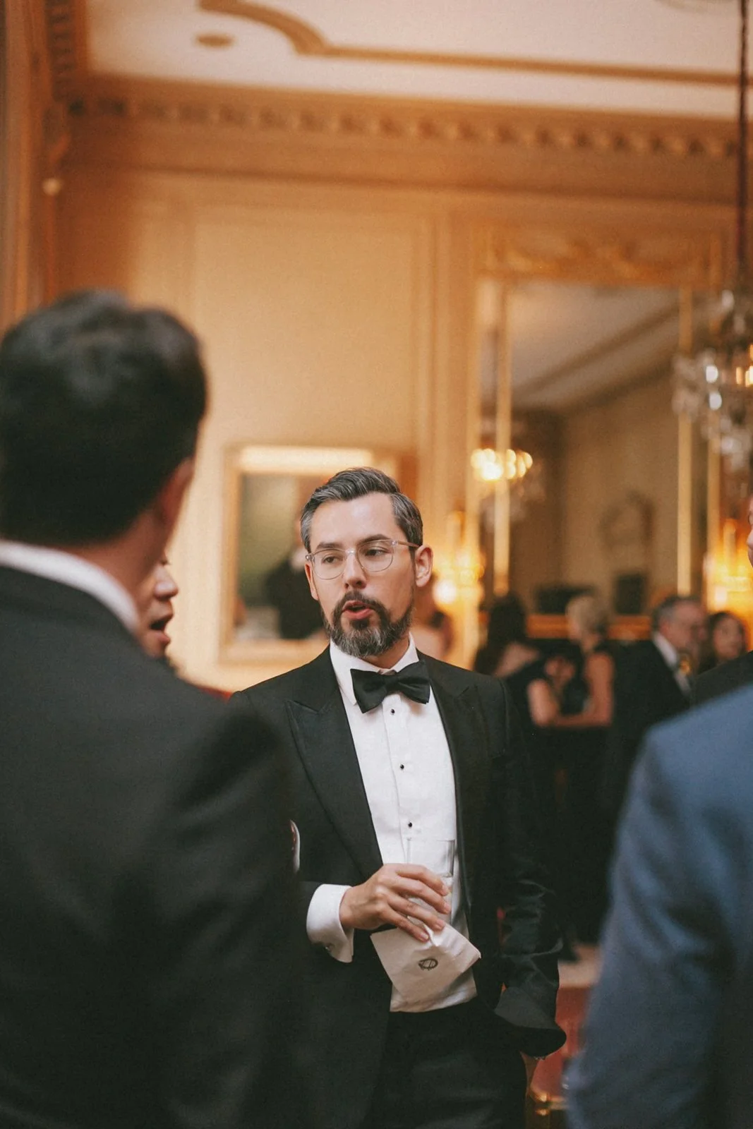 Man with glasses and beard in tuxedo holding a drink, talking to others at a formal event in an ornate room.