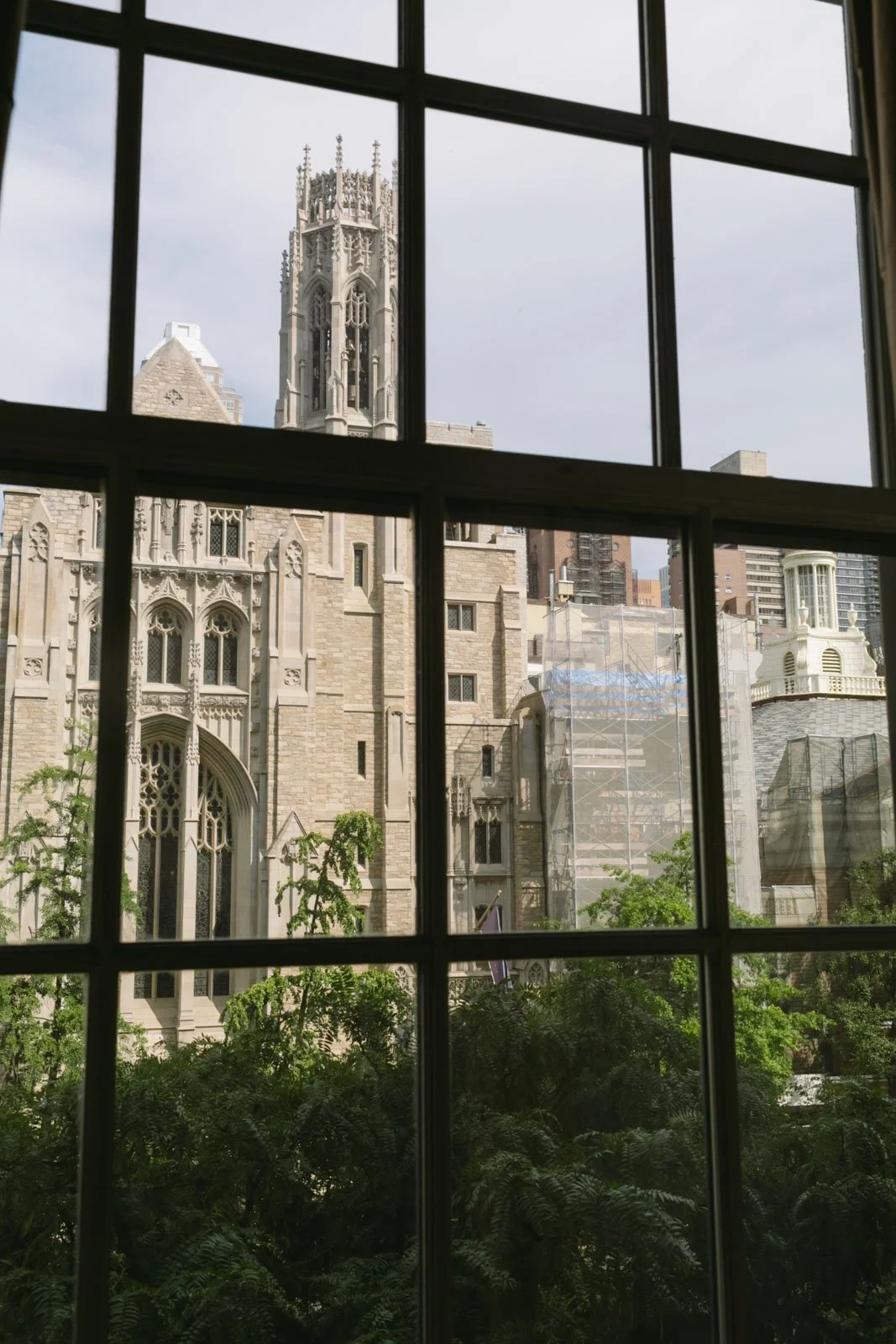 View of a historic stone church with Gothic architecture, seen through a window with multiple panes; modern buildings and greenery are visible outside.