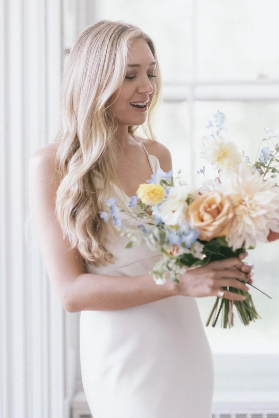 A blonde woman in a white dress holding a bouquet of pastel-colored flowers, smiling softly indoors with sheer curtains in the background.