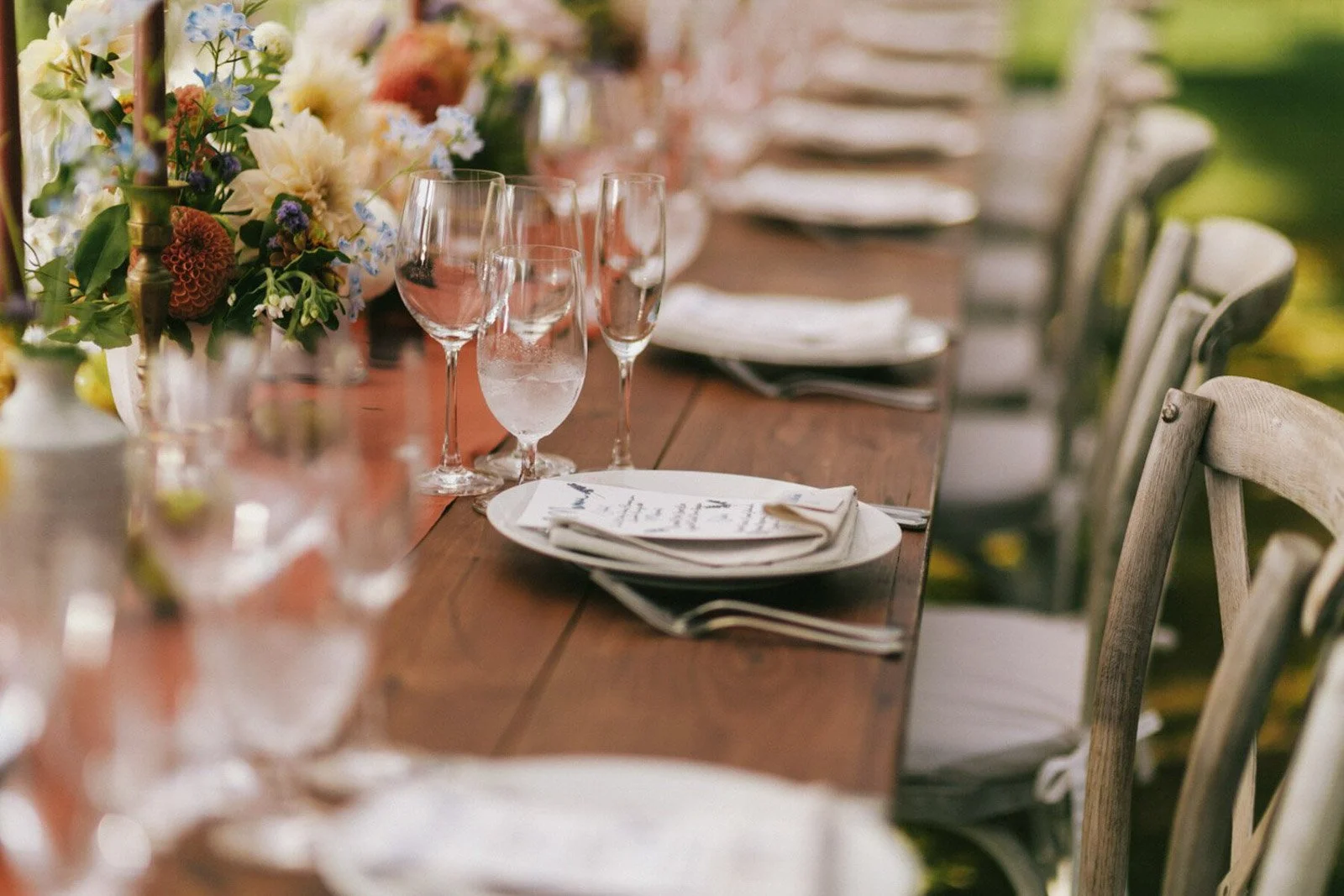 A long wooden dining table set for a meal with multiple wine glasses, plates, and napkins, decorated with a floral centerpiece featuring various flowers and greenery.