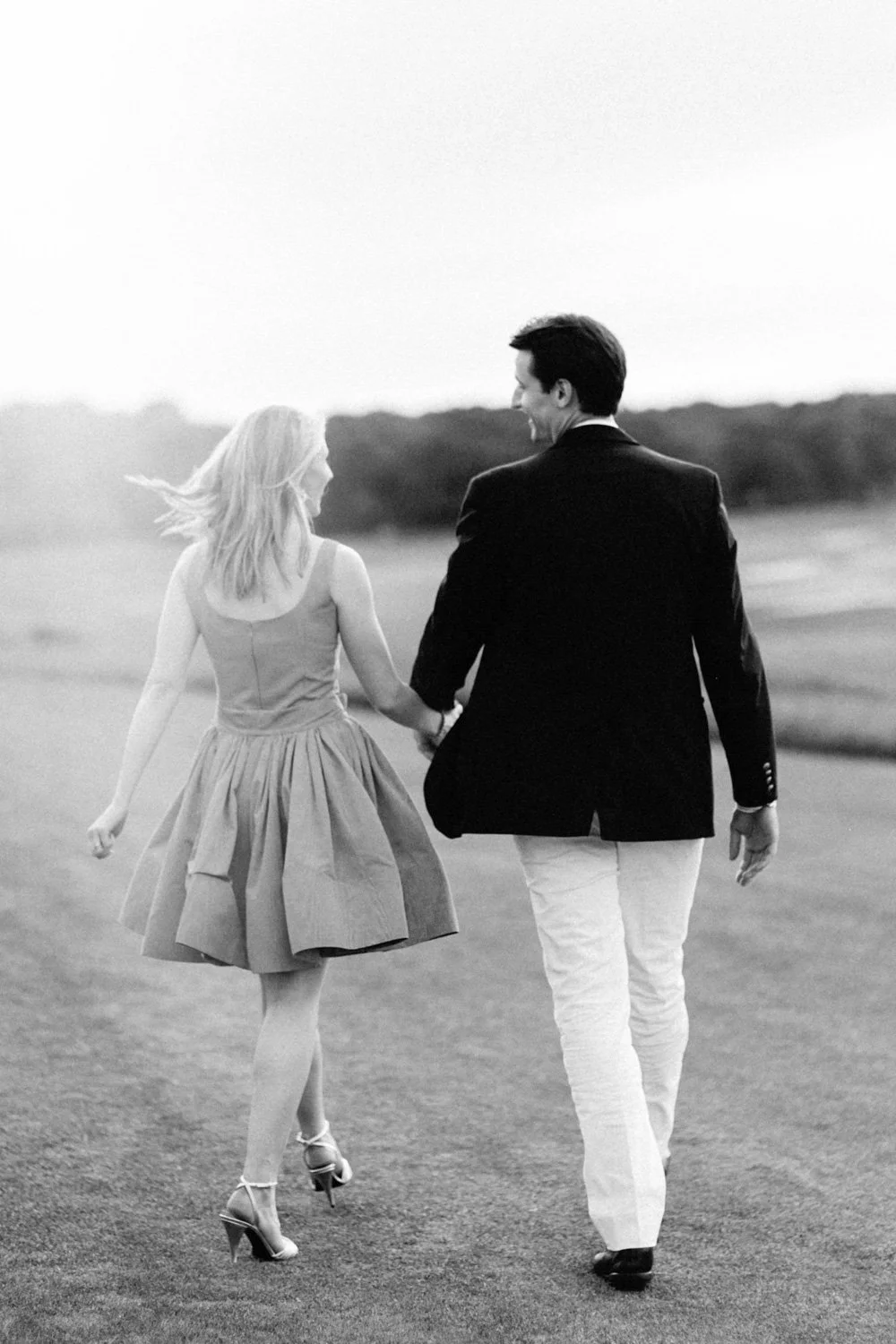 A man and woman walking hand in hand on a path in an open field, smiling and enjoying each other's company, in black and white.