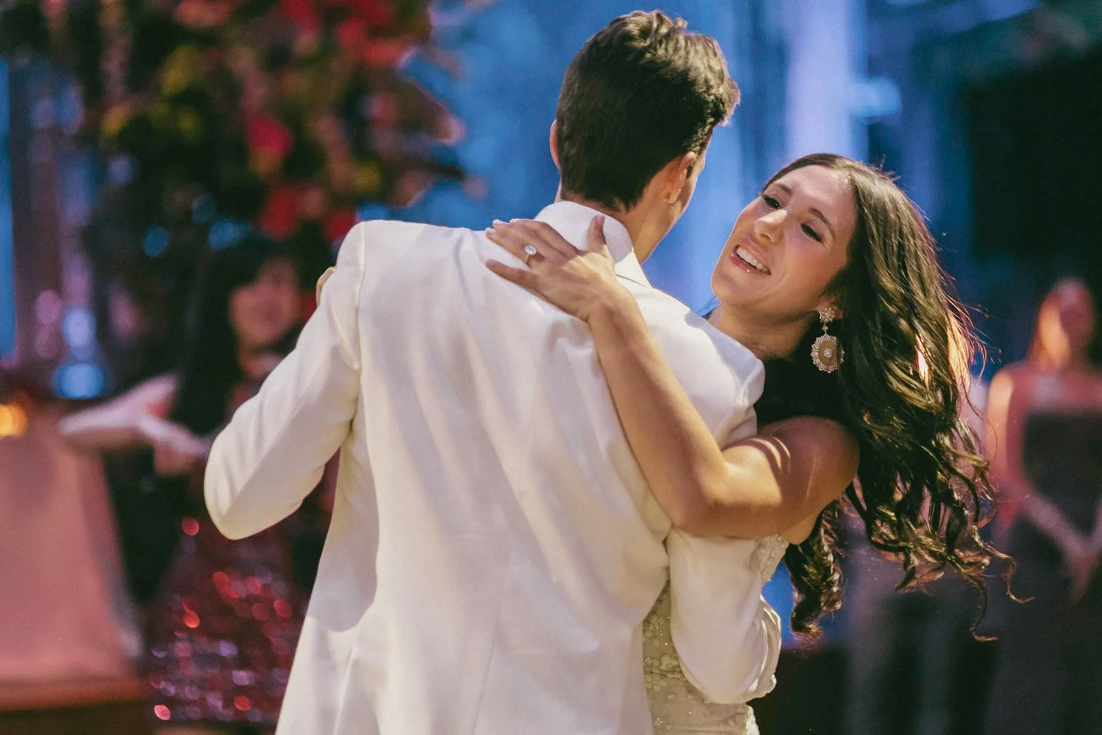 A woman and man are dancing closely at a celebration, with the woman smiling and looking happy as she holds the man, who has his back to the camera.