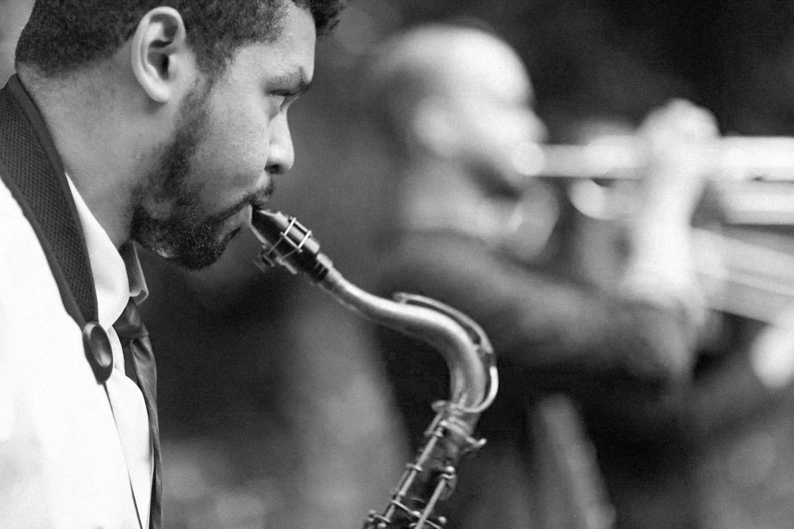 Black and white photograph of a man playing a saxophone, with focus on his face and instrument, background blurred.
