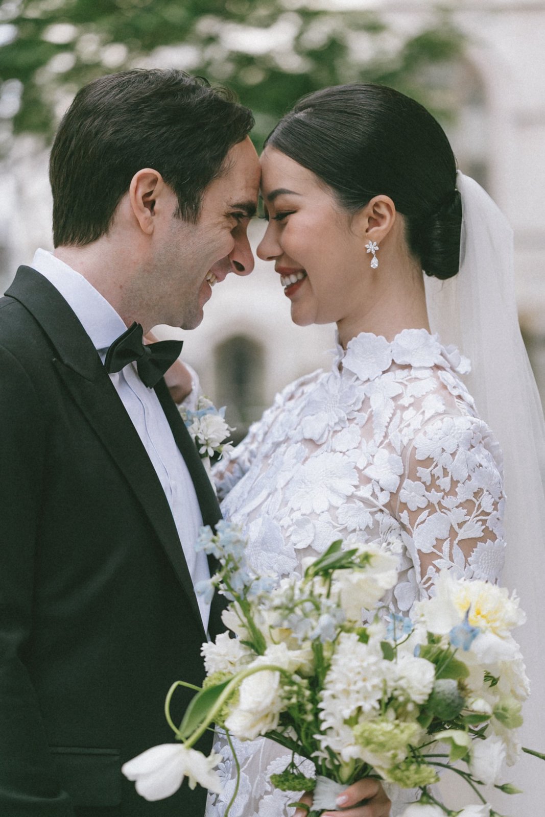A bride and groom are smiling with foreheads touching, holding a bouquet of white and light blue flowers during their wedding.