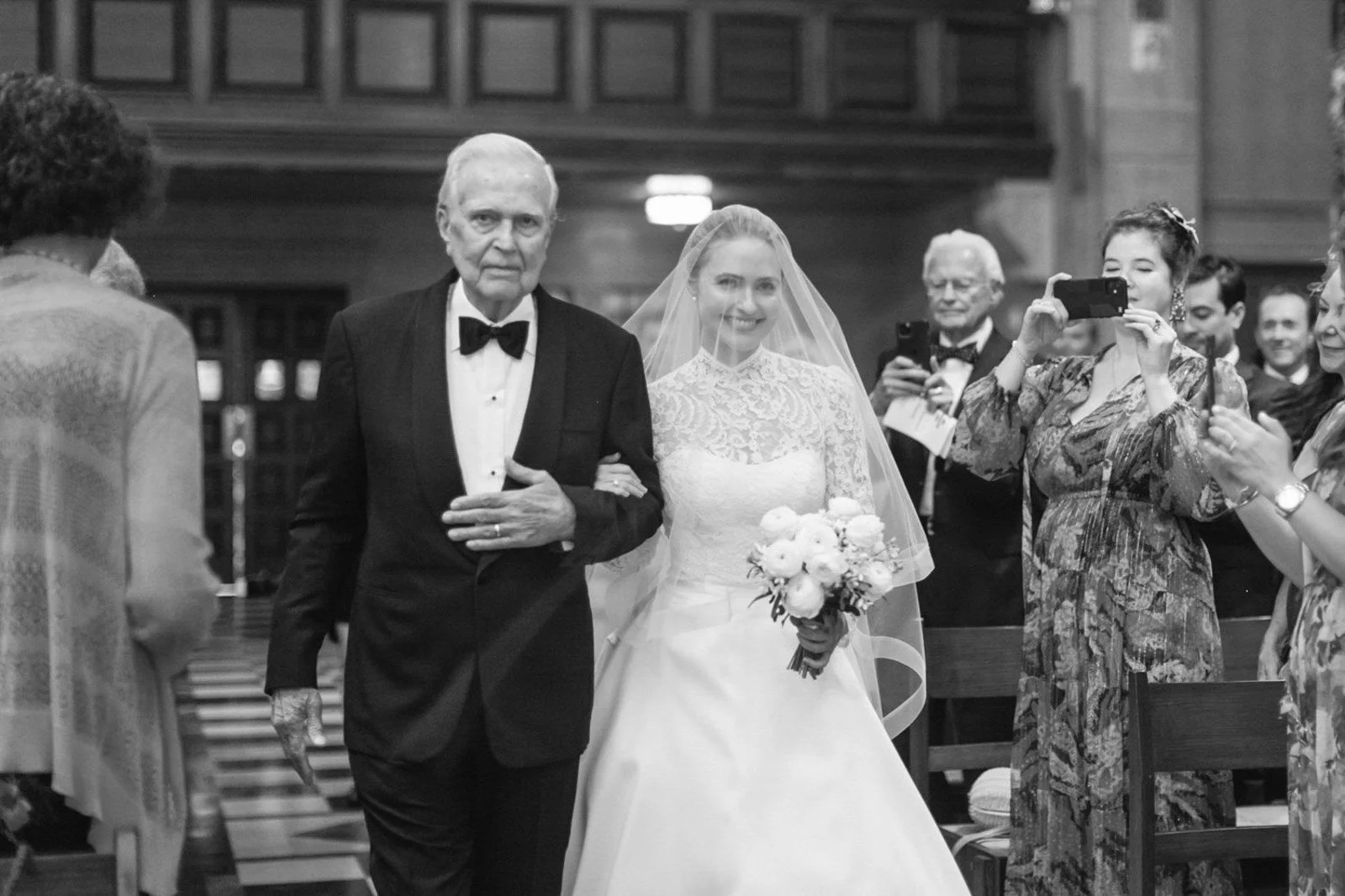 A bride walking down the aisle escorted by an elderly man at her wedding ceremony in a church, while guests take photos.