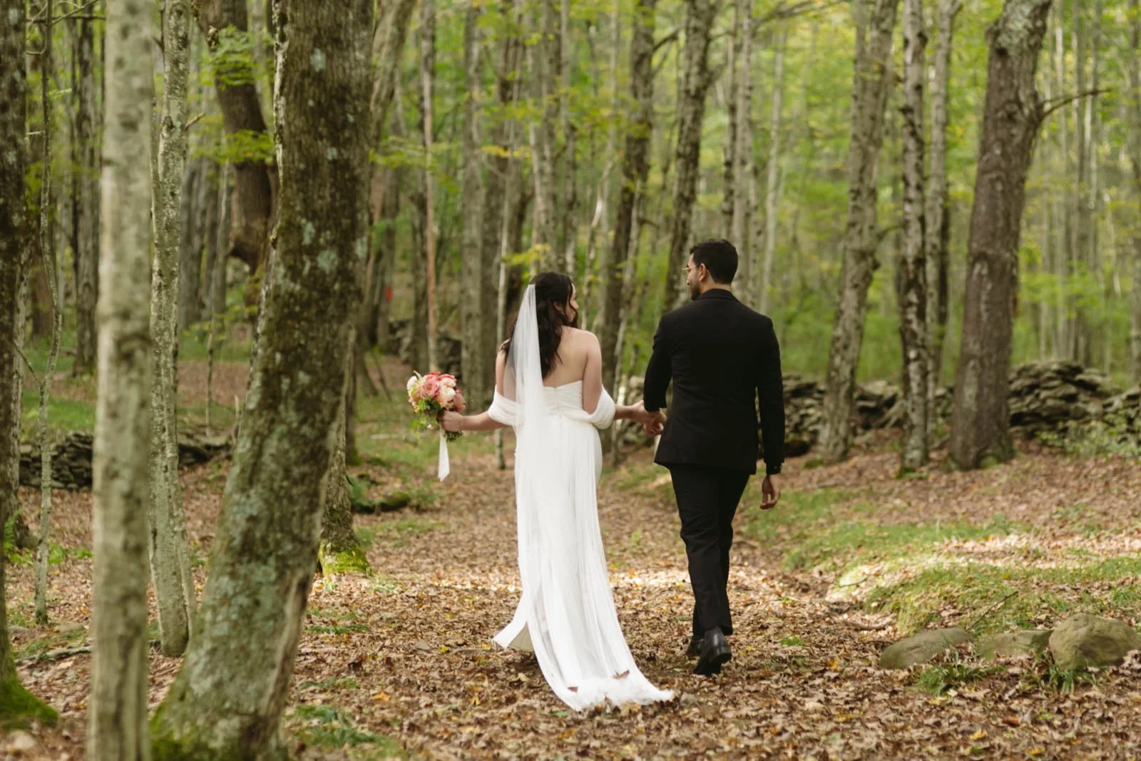 A couple dressed in wedding attire walking through a forest, with the bride holding a bouquet of flowers.