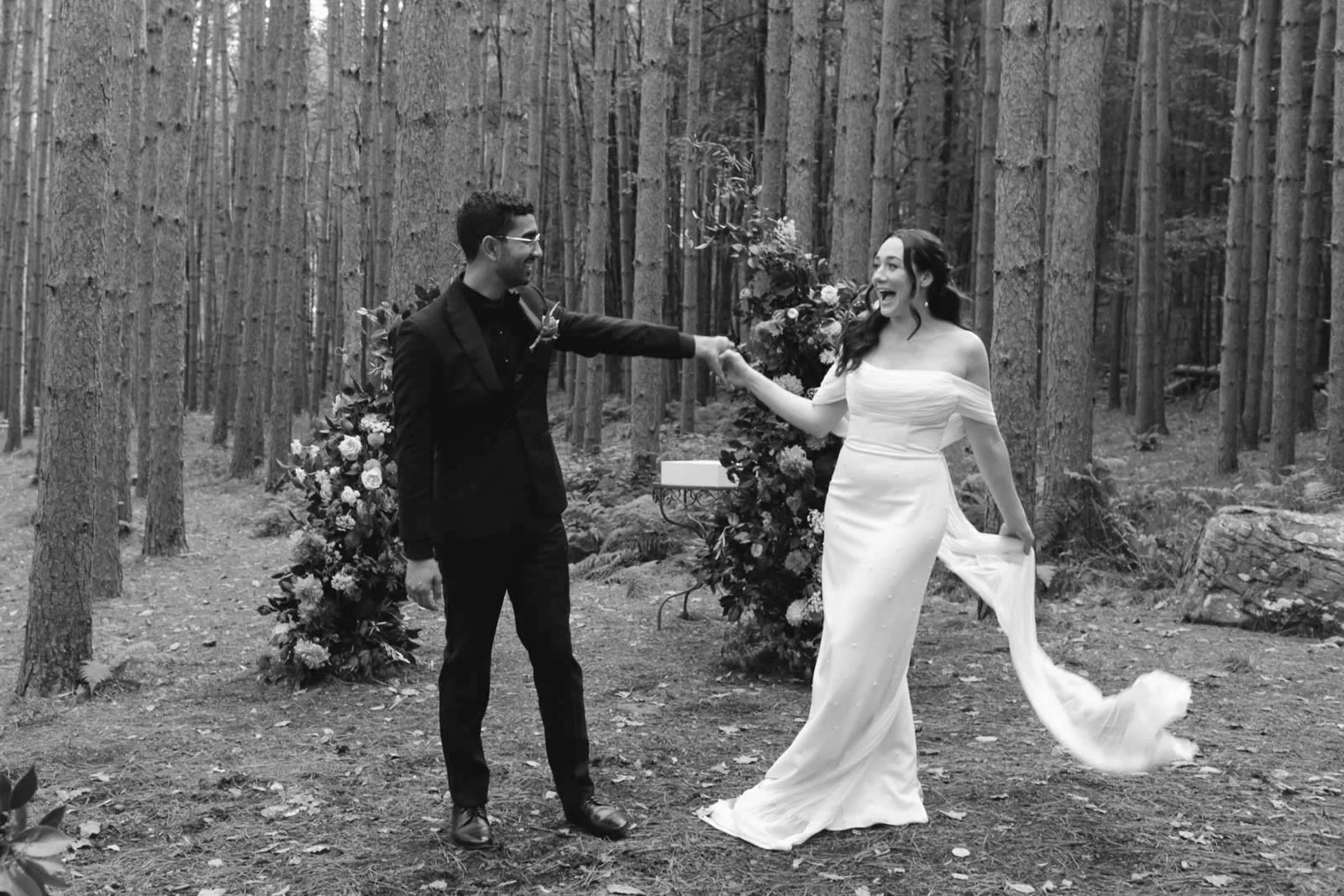 A couple getting married outdoors in a wooded area, holding hands and smiling, with wedding decorations and flowers around them in black and white.