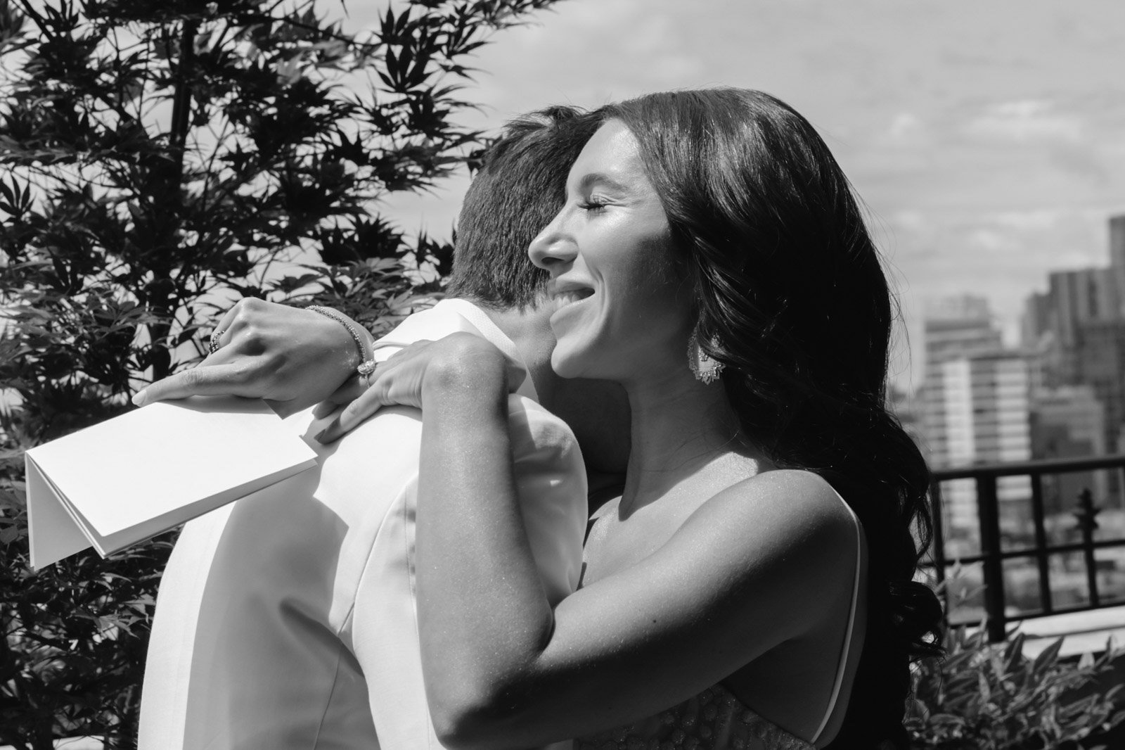 A woman embracing a man on a rooftop with city buildings in the background, smiling with her eyes closed.