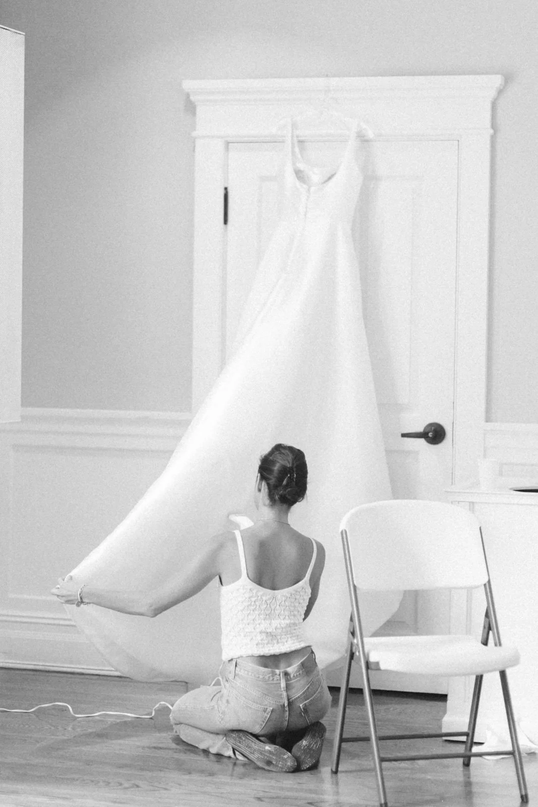 A woman kneeling on the floor holding a long piece of fabric, with a white dress hanging from the ceiling in a room with white walls and a door.