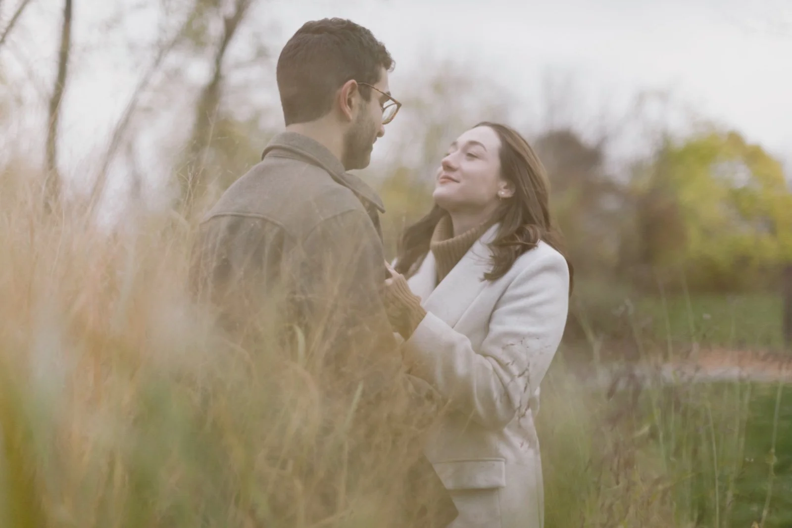 A man and woman stand close together outdoors, gazing into each other's eyes with affectionate expressions, amidst tall grass with a blurred background of trees.