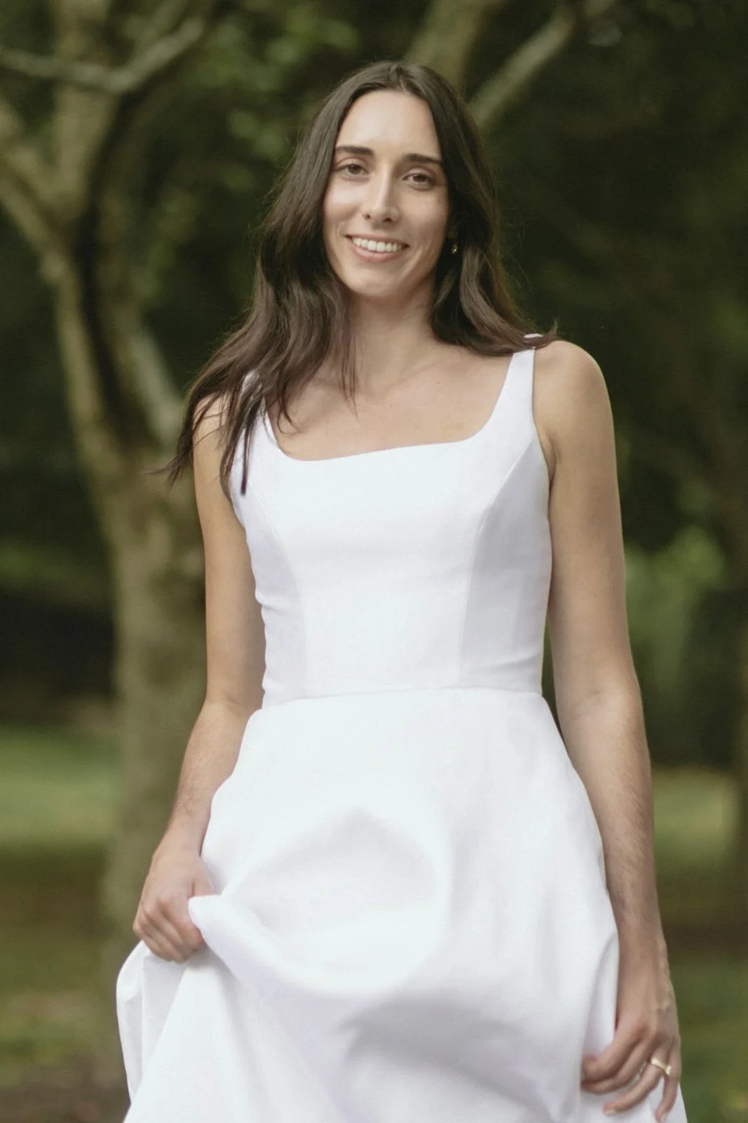 A woman with long dark hair wearing a white dress, standing outdoors near trees, smiling at the camera.